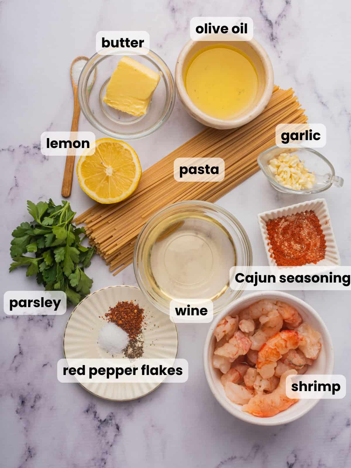 Ingredients needed to make spicy shrimp scampi on a marble countertop include Cajun seasoning, pasta, white wine, garlic, butter, and olive oil.