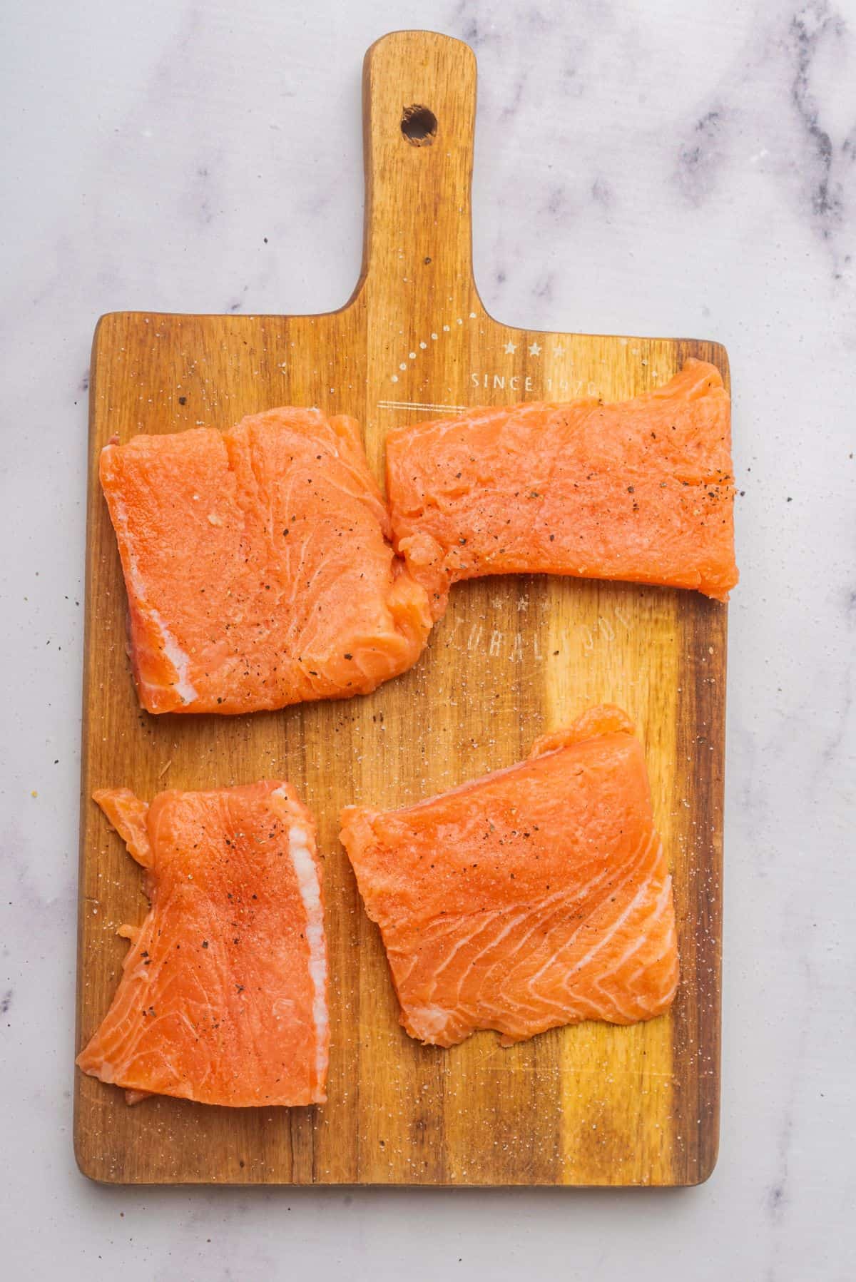 Four skinless salmon fillets with salt and pepper are on a wooden cutting board.