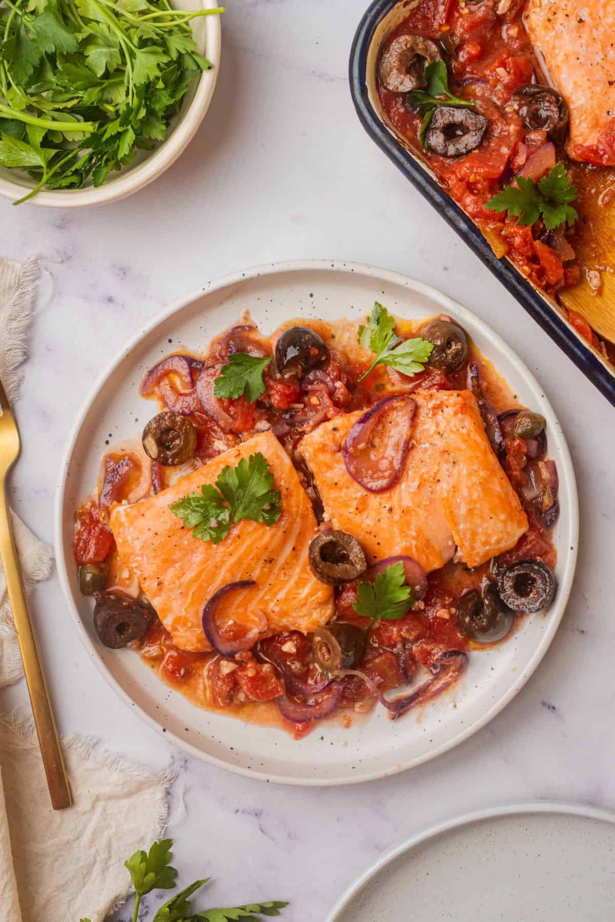 Two fillets of salmon puttanesca are on a plate with a tomato sauce made with olives and capers.