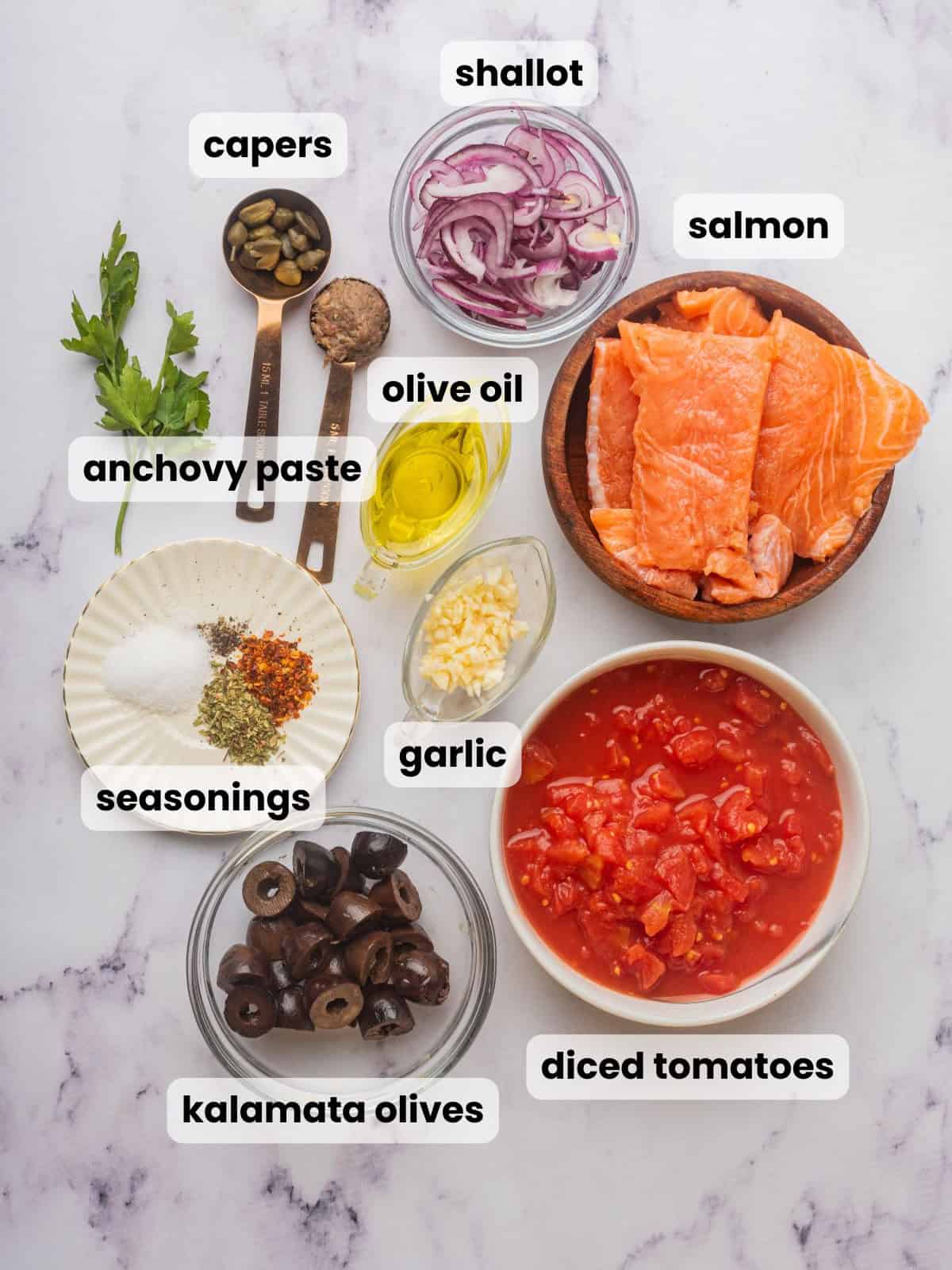 Ingredients needed to make fish puttanesca are on a countertop: salmon, tomatoes, capers, anchovy paste, olives, and seasonings.