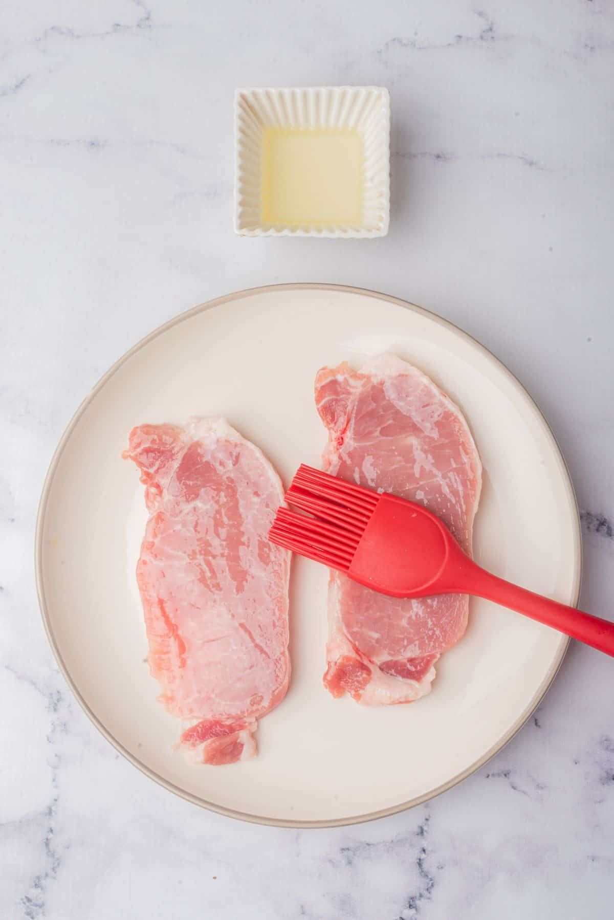 Thin boneless pork chops on a plate next to a bowl of olive oil and a pastry brush.