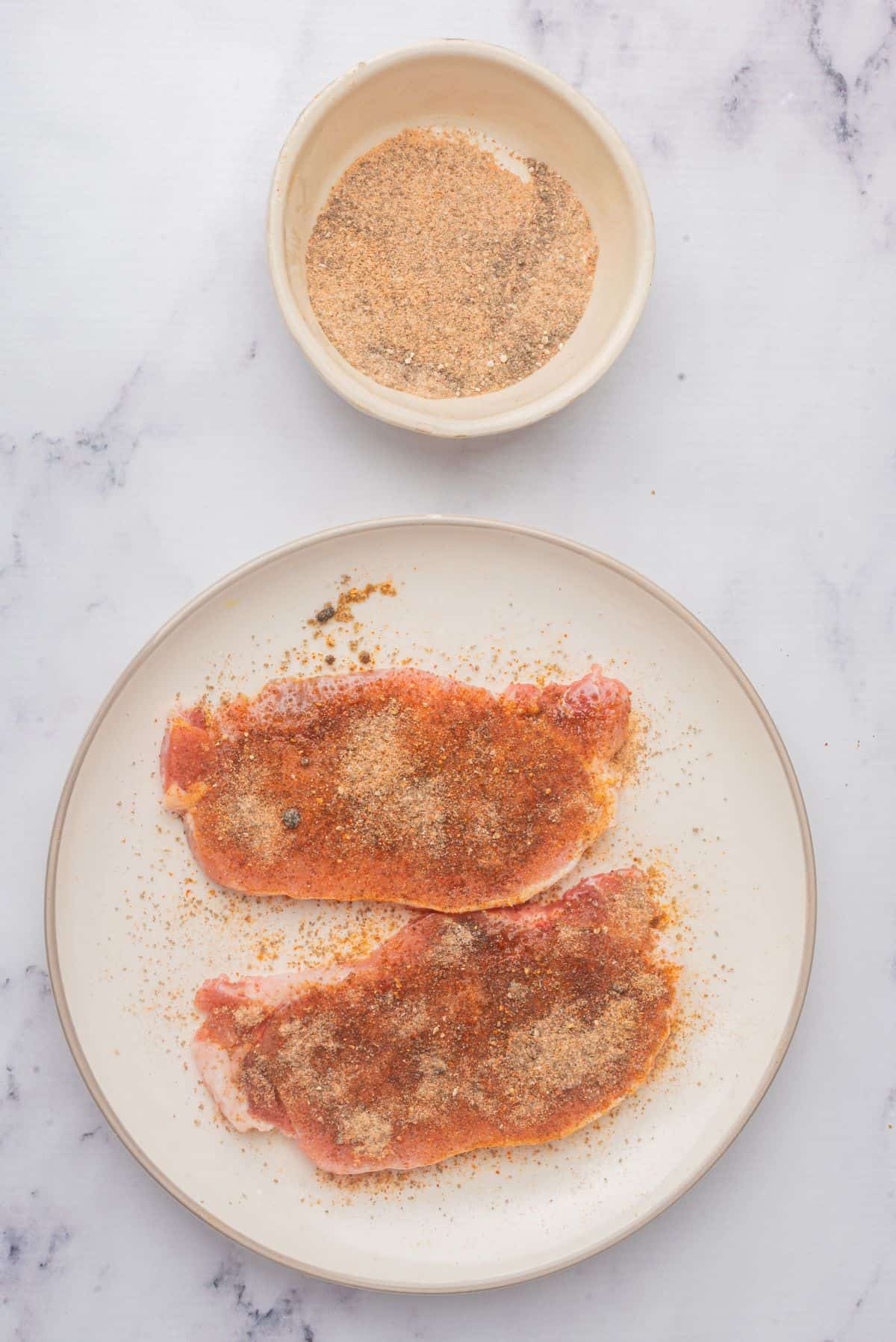 Seasoned thin boneless pork chops on a plate next to a bowl of seasonings.