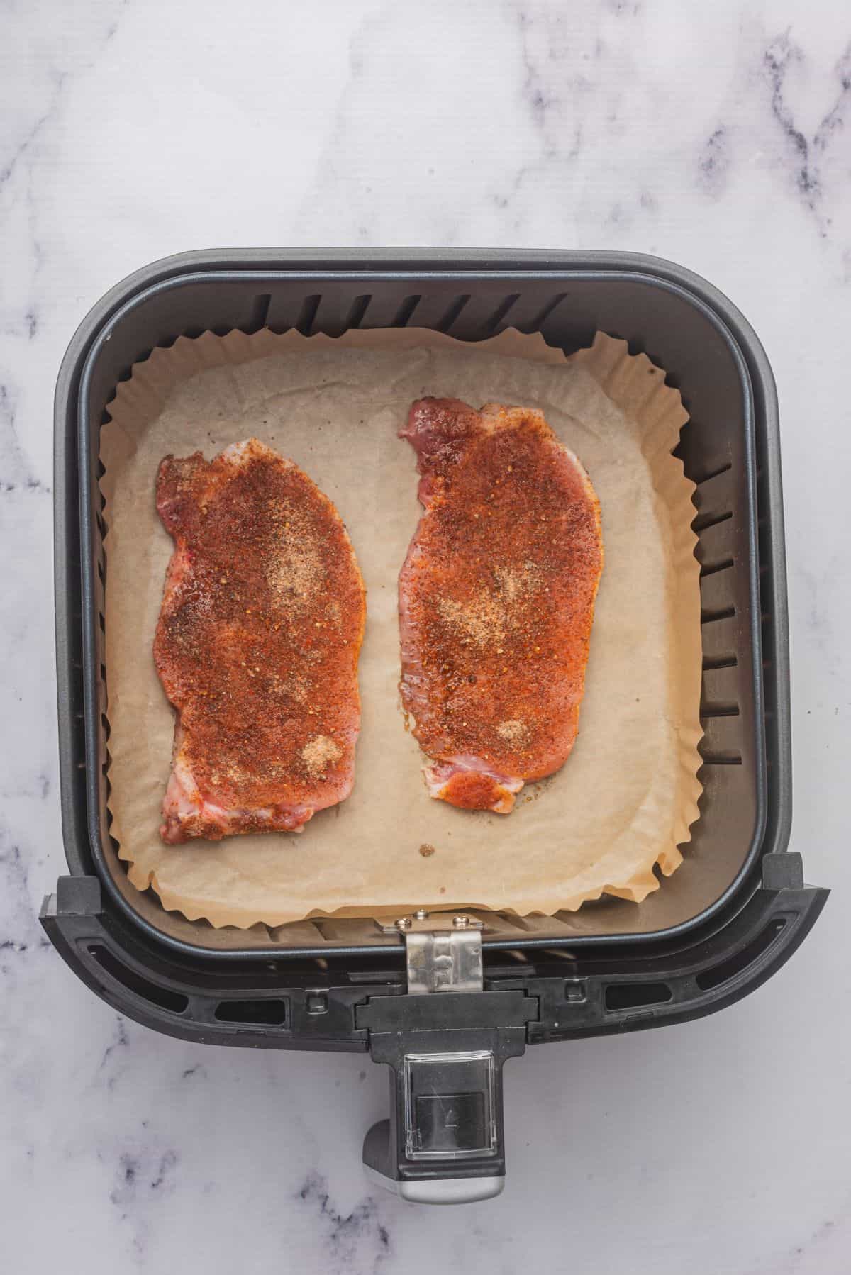 Two raw and seasoned thin pork chops in air fryer basket.