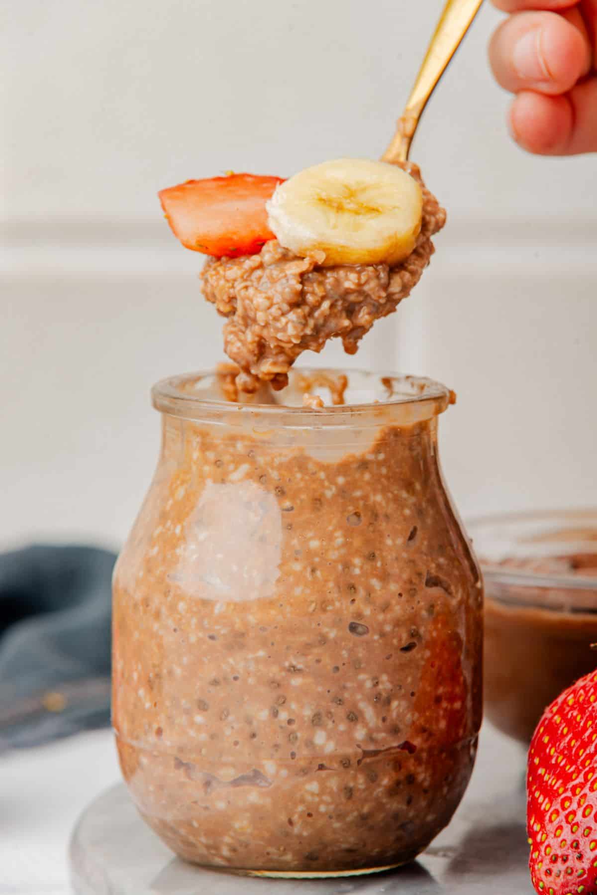 Nutella overnight oats in a jar with a spoon scooping out a bite with a banana and strawberry.