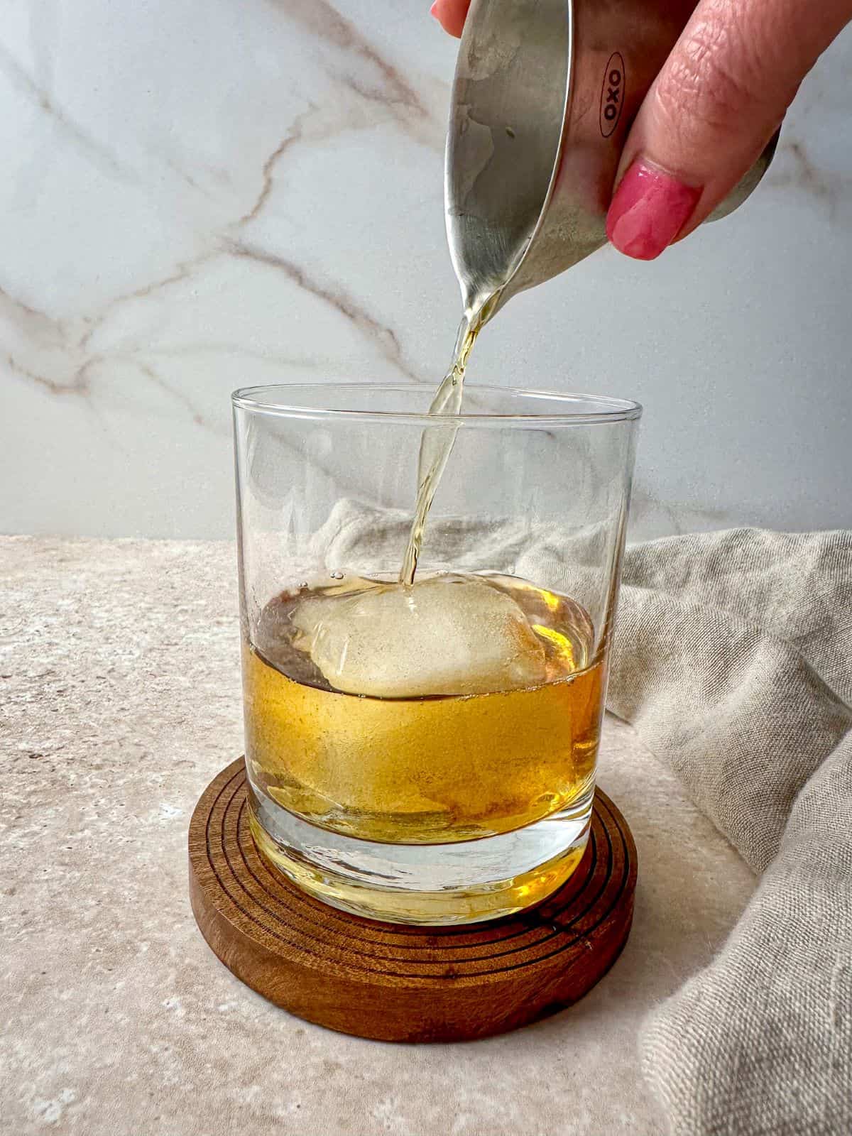 A hand pouring bourbon over a large ice cube in a rocks glass.