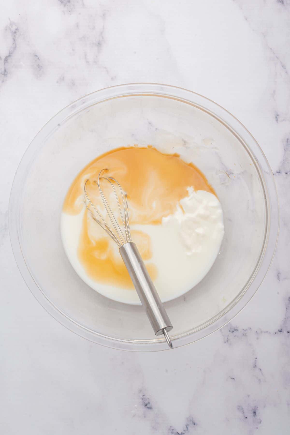 A whisk in a glass bowl with vanilla almond milk, maple syrup, vanilla Greek yogurt, and vanilla extract.