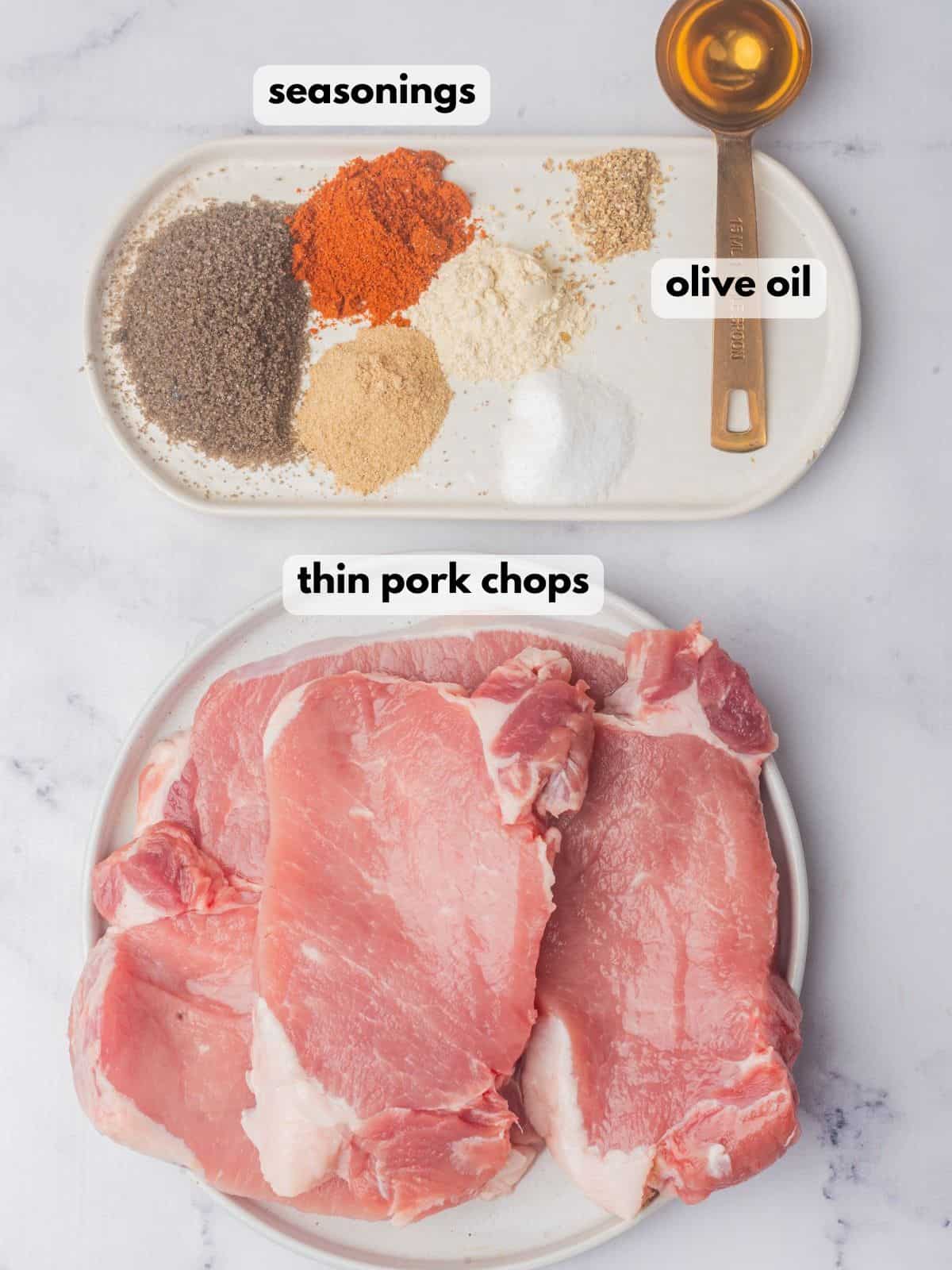 Thin cut pork chops, a tablespoon of olive oil, and a bowl of seasonings on a marble countertop.