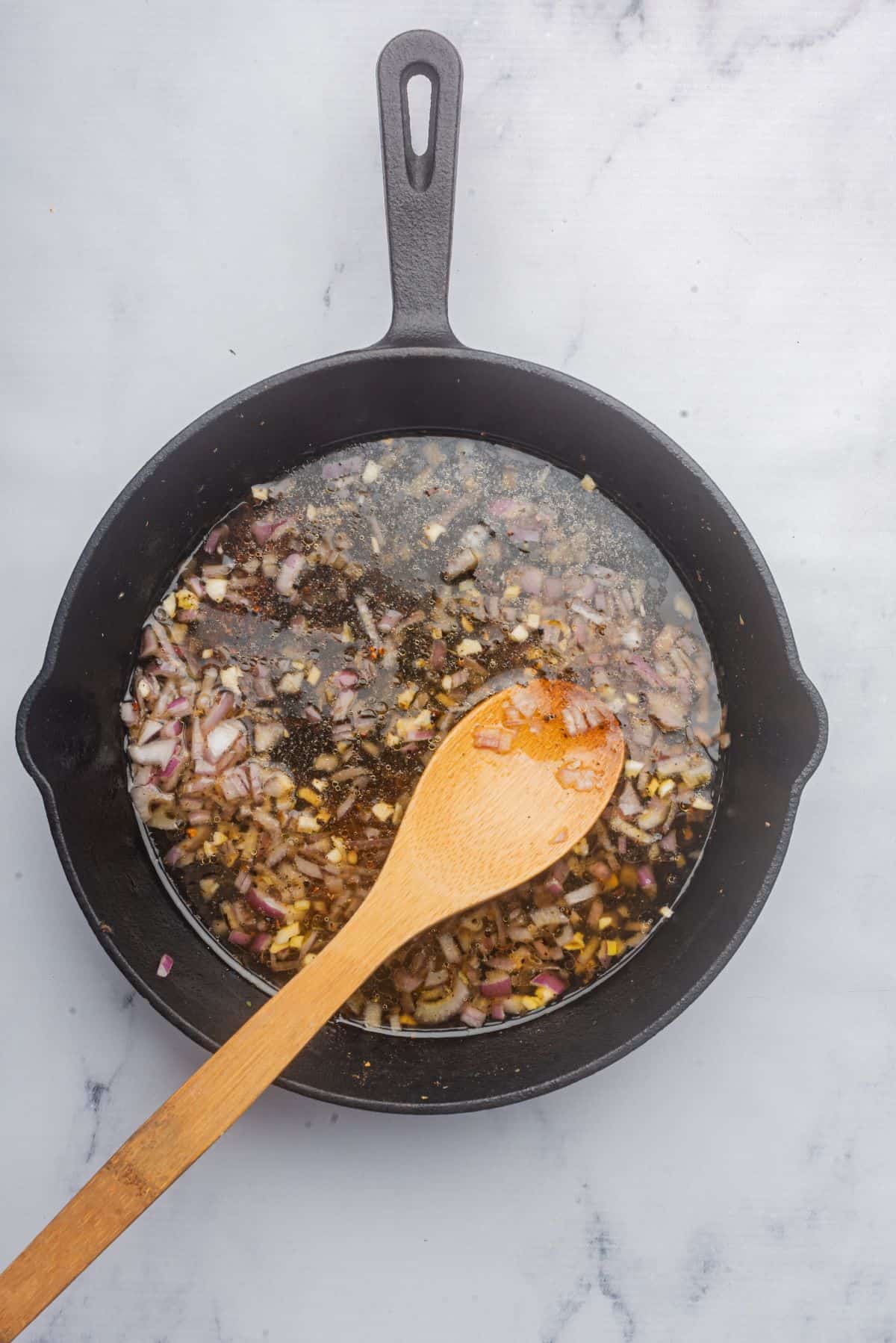 Garlic, shallots, and chicken broth in a cast iron skillet with a wooden spoon.