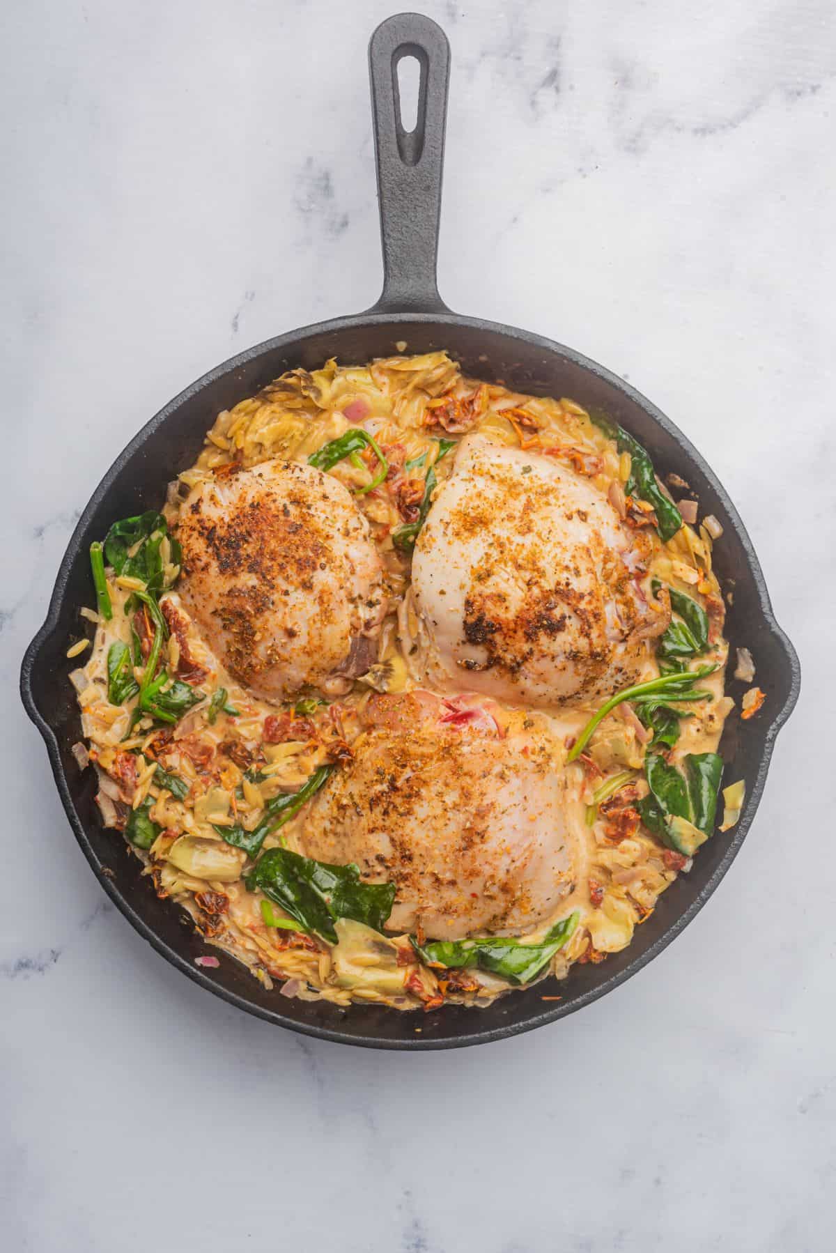 Chicken and orzo in a skillet with ingredients ready to be baked.