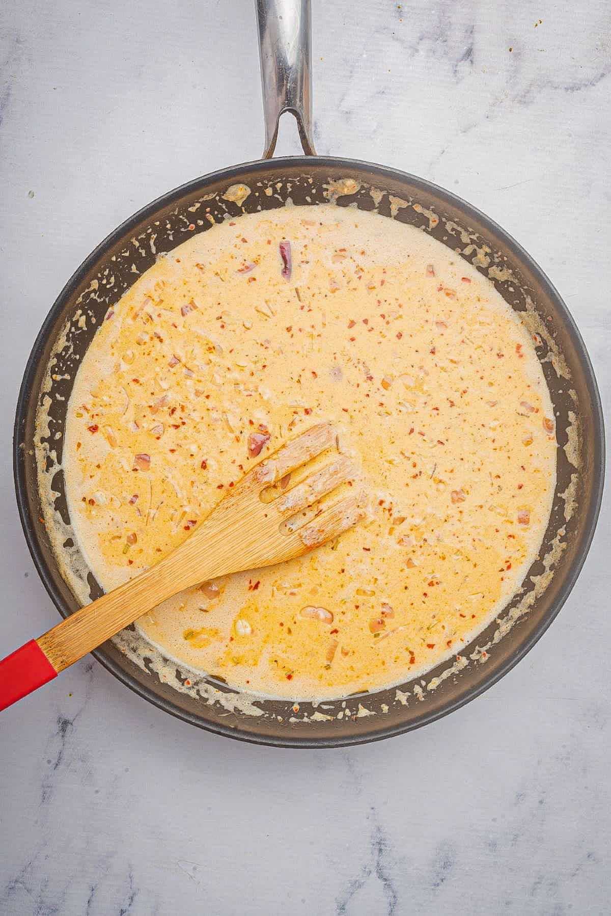 Cajun Alfredo sauce recipe cooking in a large skillet with a wooden spoon.