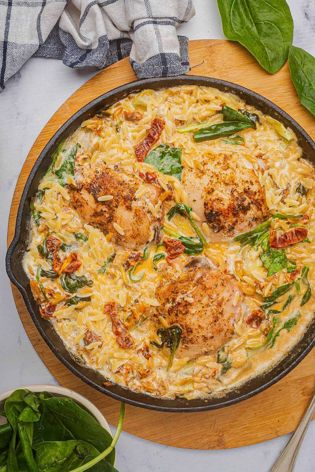 Baked chicken and orzo pasta in a skillet with spinach, artichoke hearts, and sun-dried tomatoes.