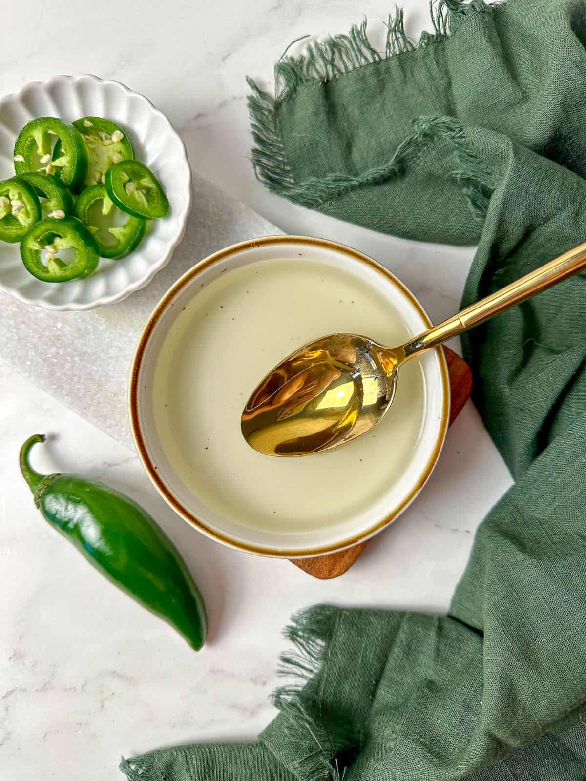 Homemade spicy jalapeno simple syrup in a bowl with a spoon surrounded by fresh jalapenos.