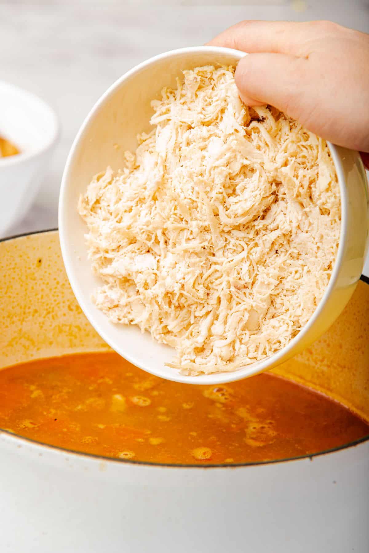 A hand adding shredded rotisserie chicken into a pot of tortilla soup.