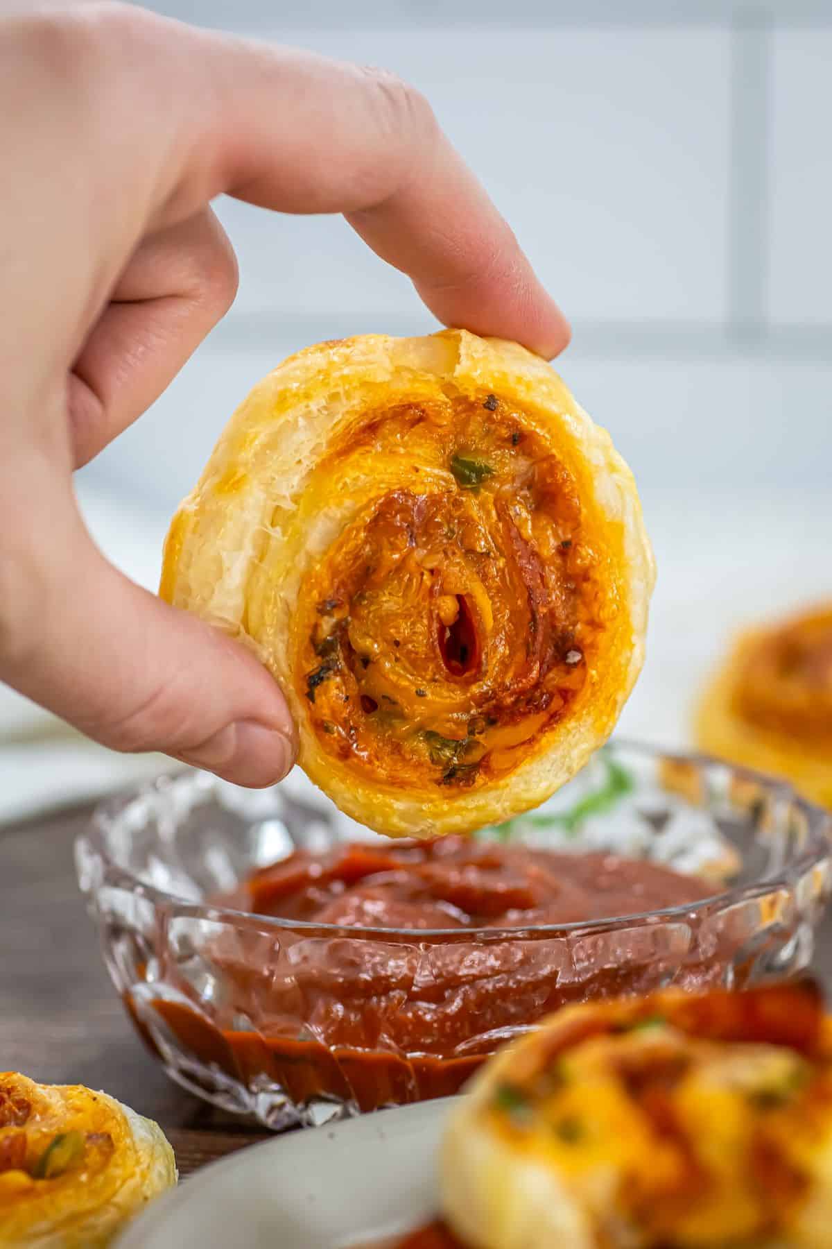 A hand dipping a pepperoni pizza pinwheel in marinara sauce.