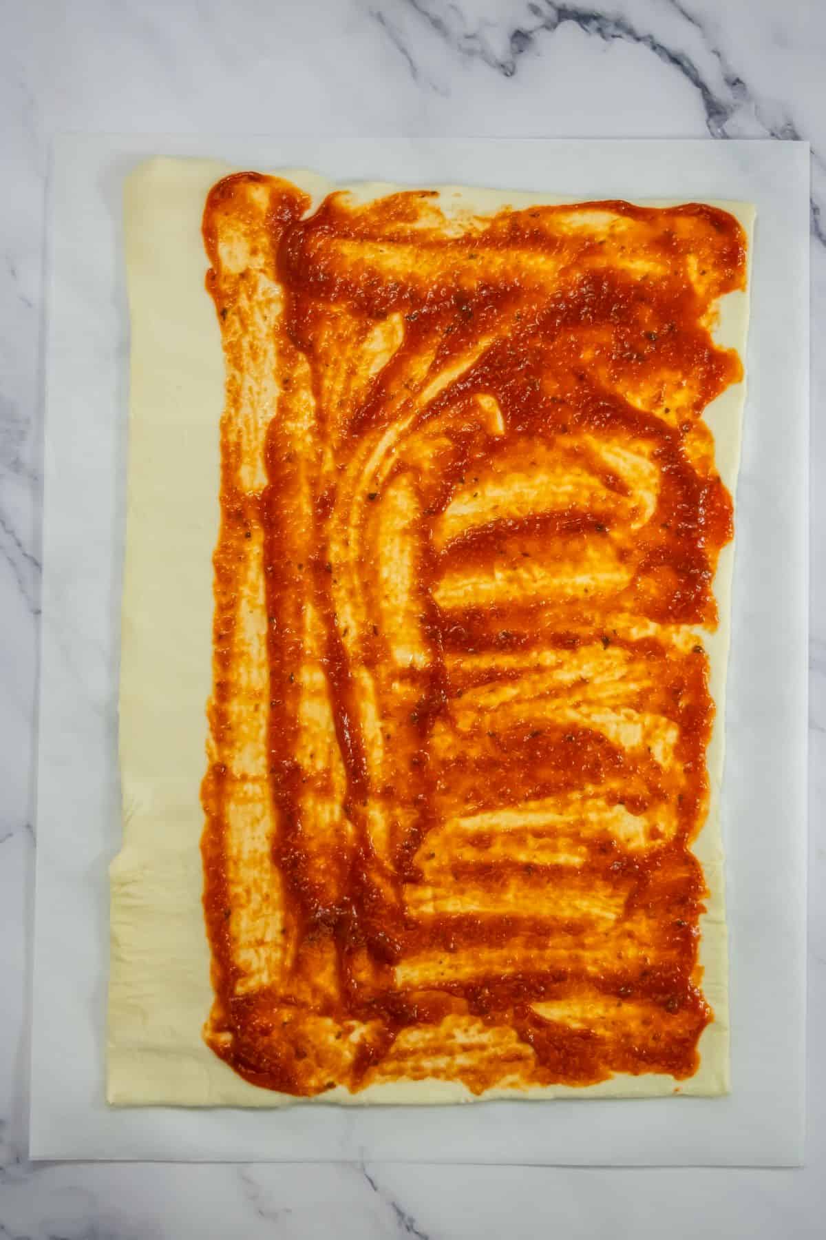 Puff pastry sheet rolled out flat with marinara sauce on it.