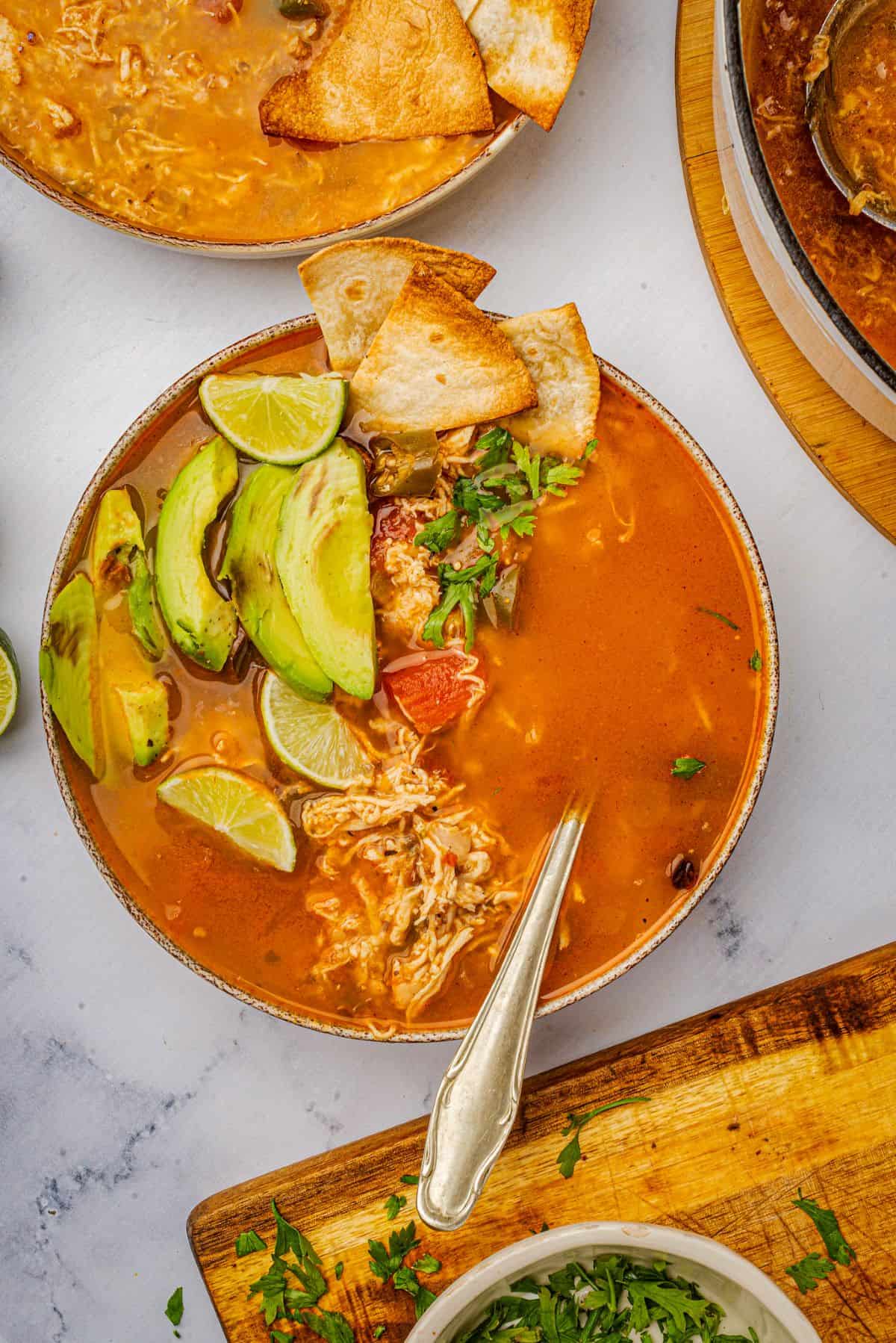 A bowl of tortilla soup with rotisserie chicken topped with avocado, cilantro, lime wedge, and tortilla chips.