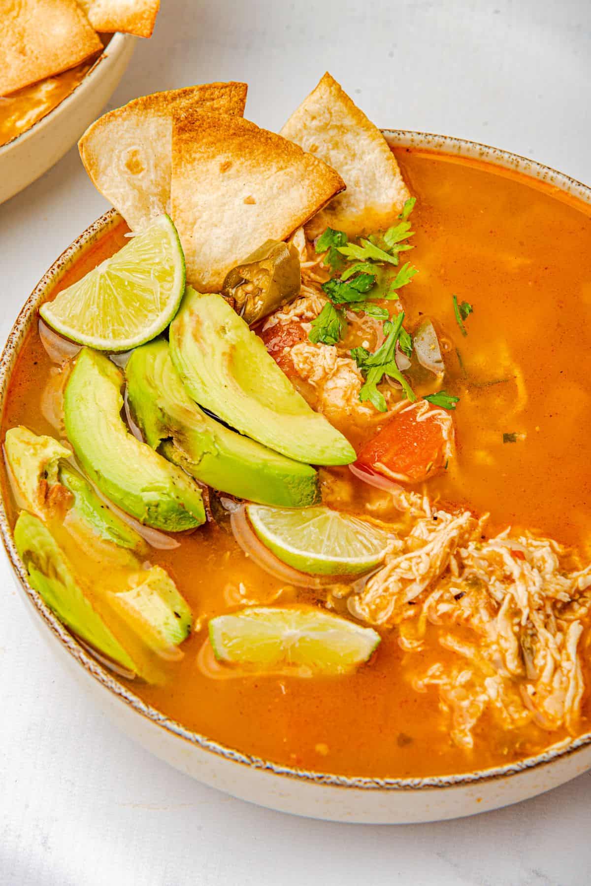 A bowl of homemade rotisserie chicken tortilla soup topped with avocado, cilantro, lime wedge, and tortilla chips.