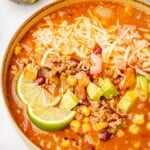 Beef enchilada soup made with ground beef and red enchilada sauce in a bowl with corn, black beans, cheese, avocado slices, and lime wedge.