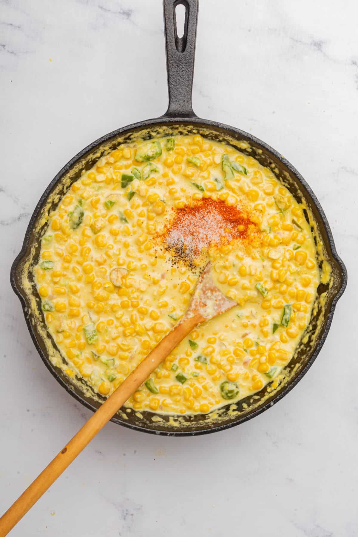 A wooden spoon and seasonings added to the skillet of cream corn with jalapenos.