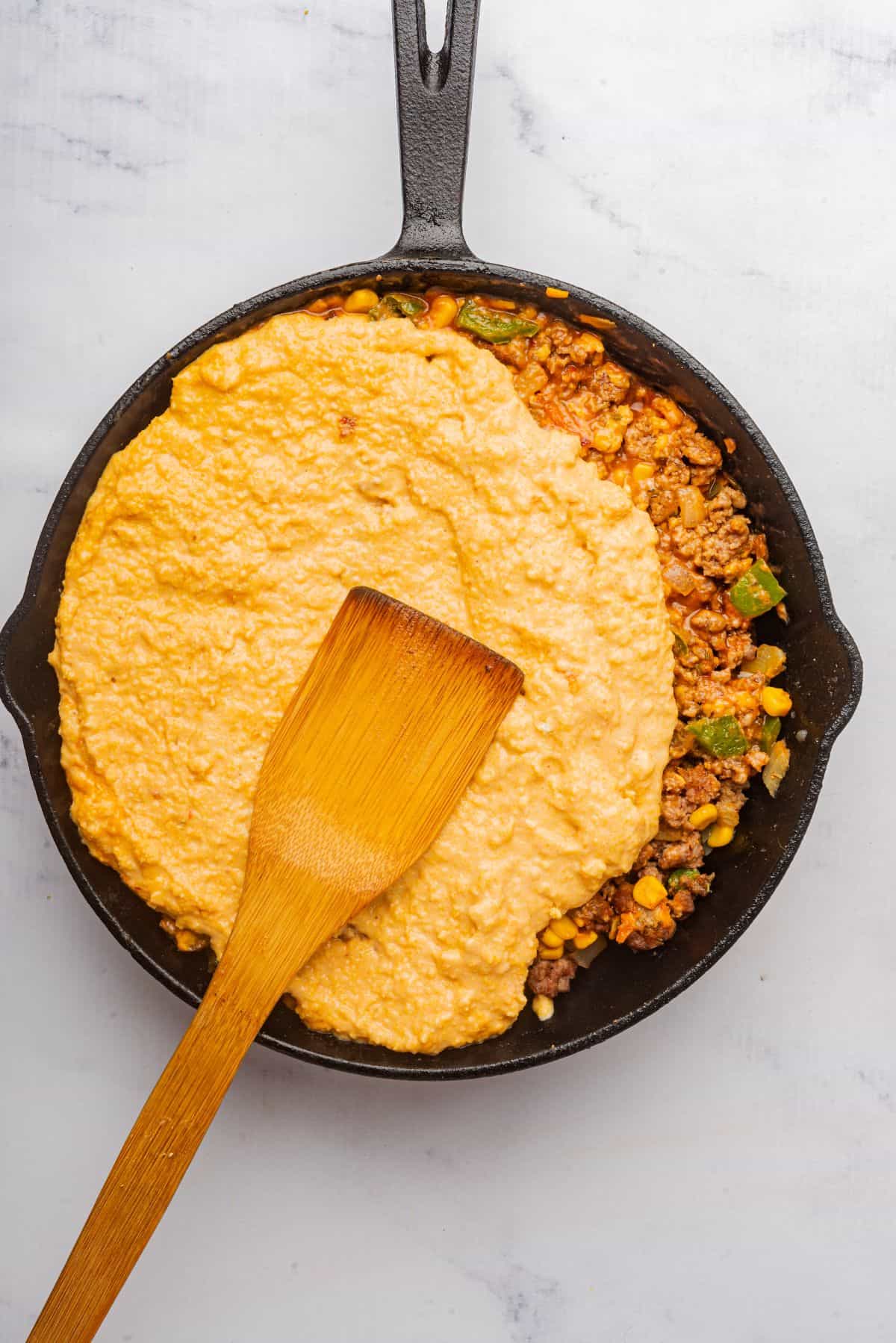 A spatula spreading cornbread crust mixture over the top of the ground beef meat mixture.