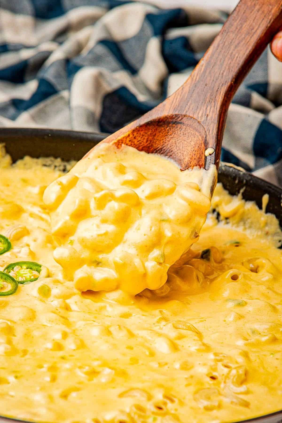 Jalapeno mac and cheese falling off a wooden spoon into a skillet.