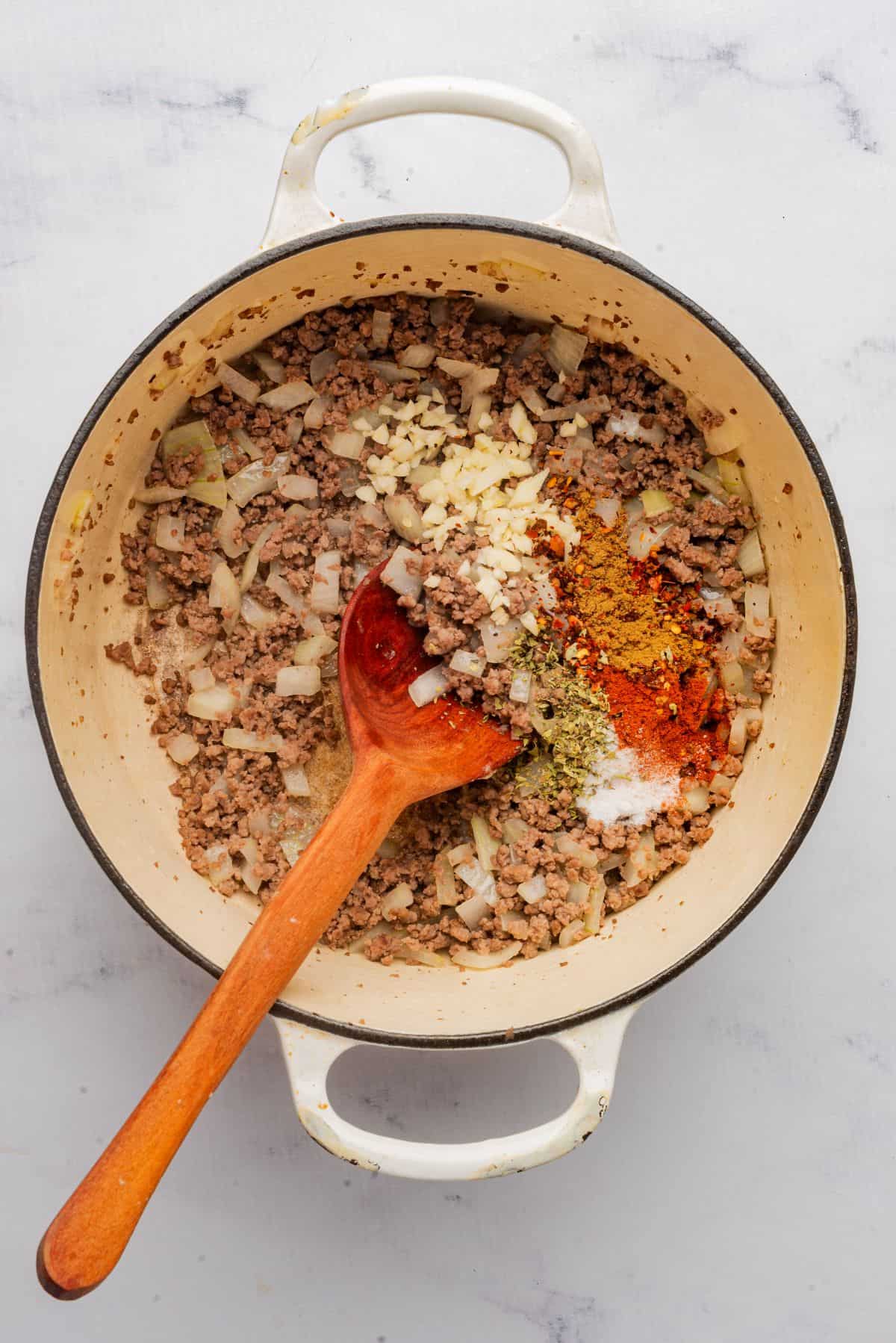 Ground beef, onions, and spices sauteing in a Dutch oven with a wooden spoon.