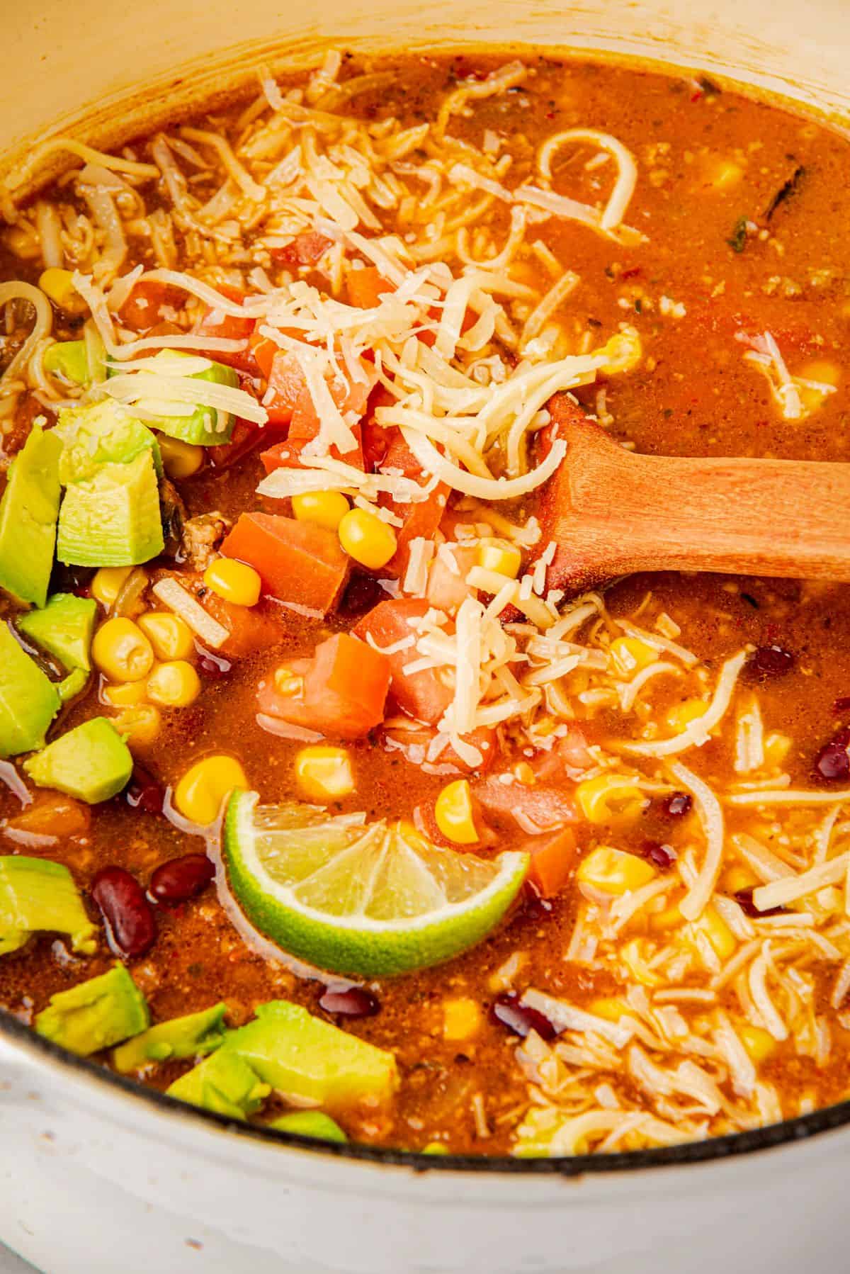 Best beef enchilada soup in a pot with cheese, diced tomatoes, corn, beans, avocado slices, a wooden spoon.