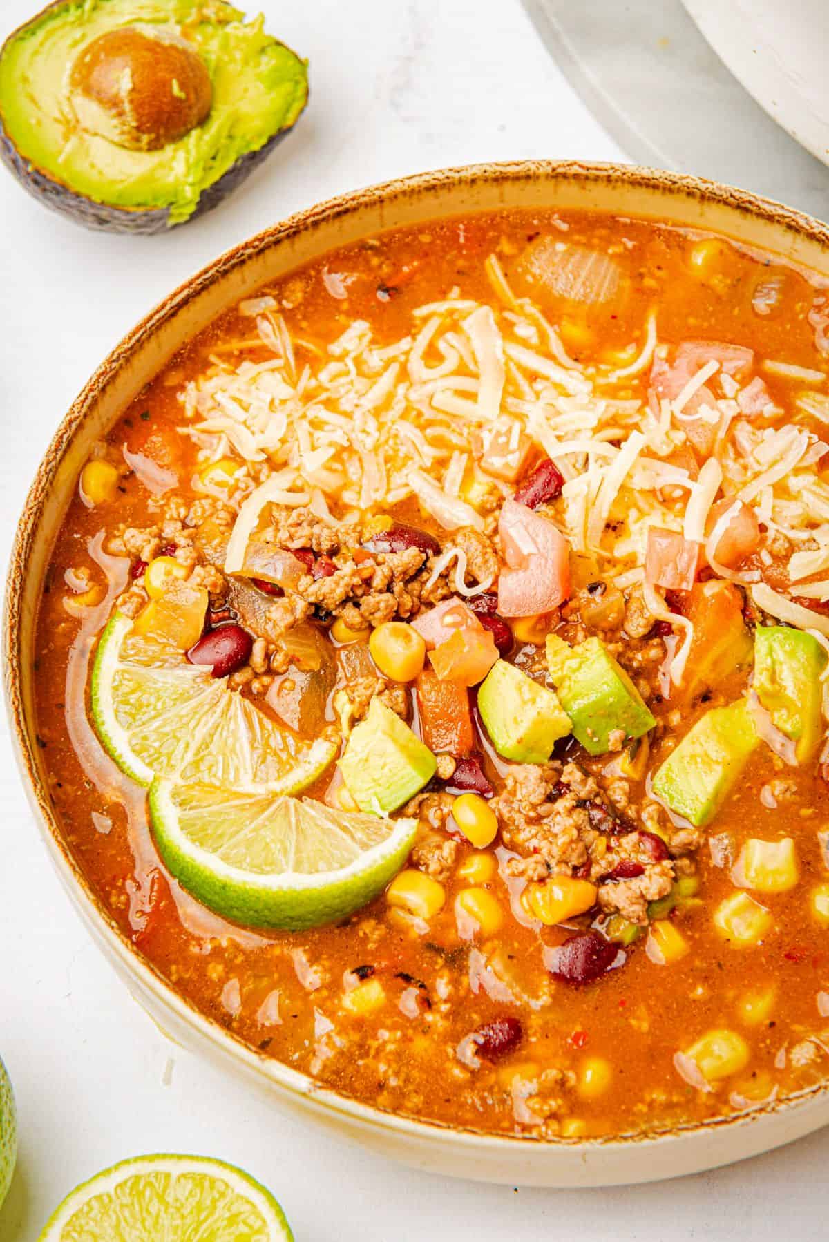 Beef enchilada soup made with ground beef and red enchilada sauce in a bowl with corn, beans, cheese, avocado slices, and lime wedge.