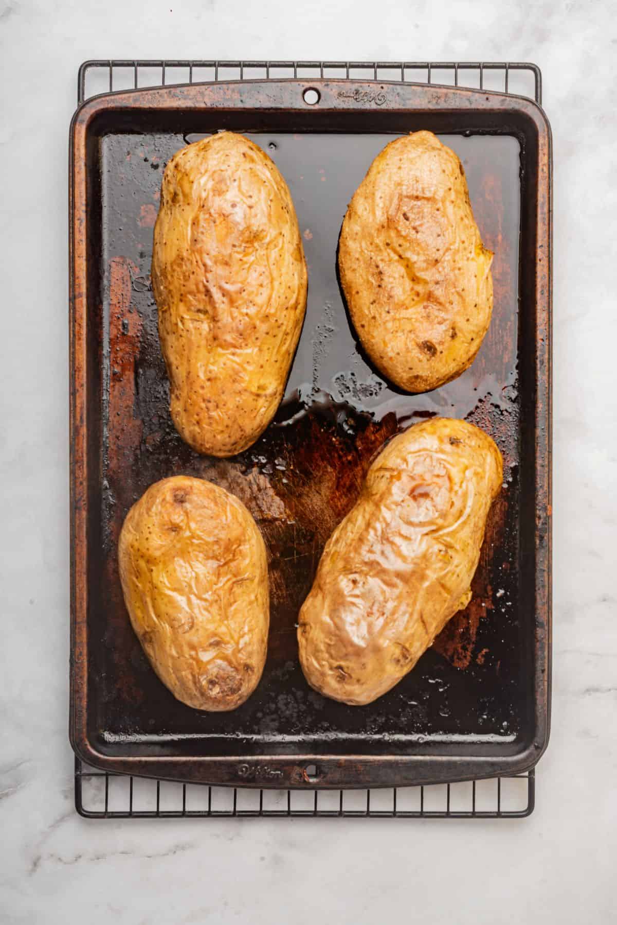 Four baked potatoes on a sheet pan with crispy skin.