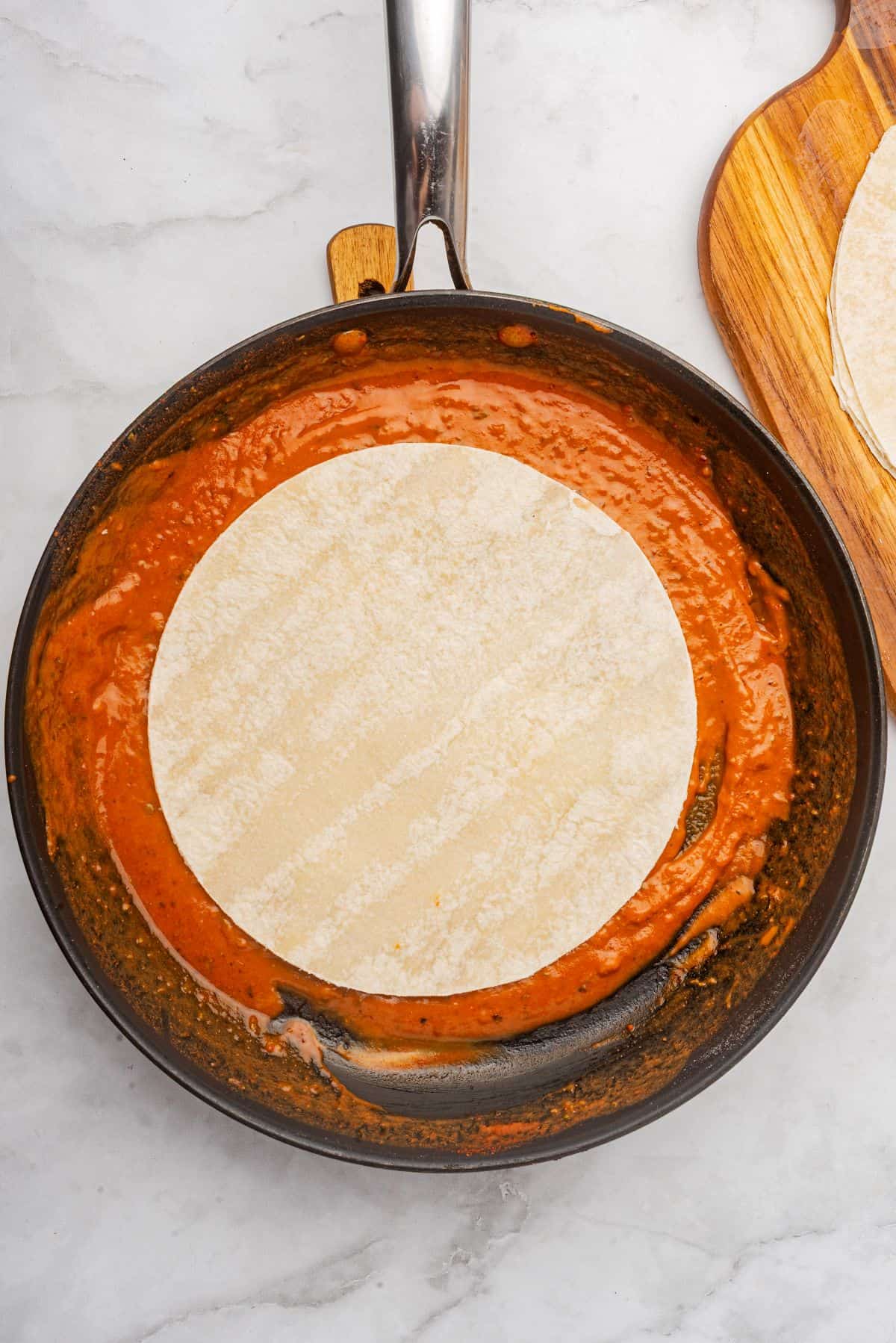 A tortilla dipped into a skillet of red enchilada sauce.