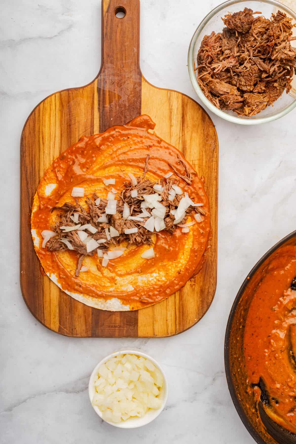 A tortilla on a cutting board coated in red enchilada sauce and filled with shredded brisket and white onions.