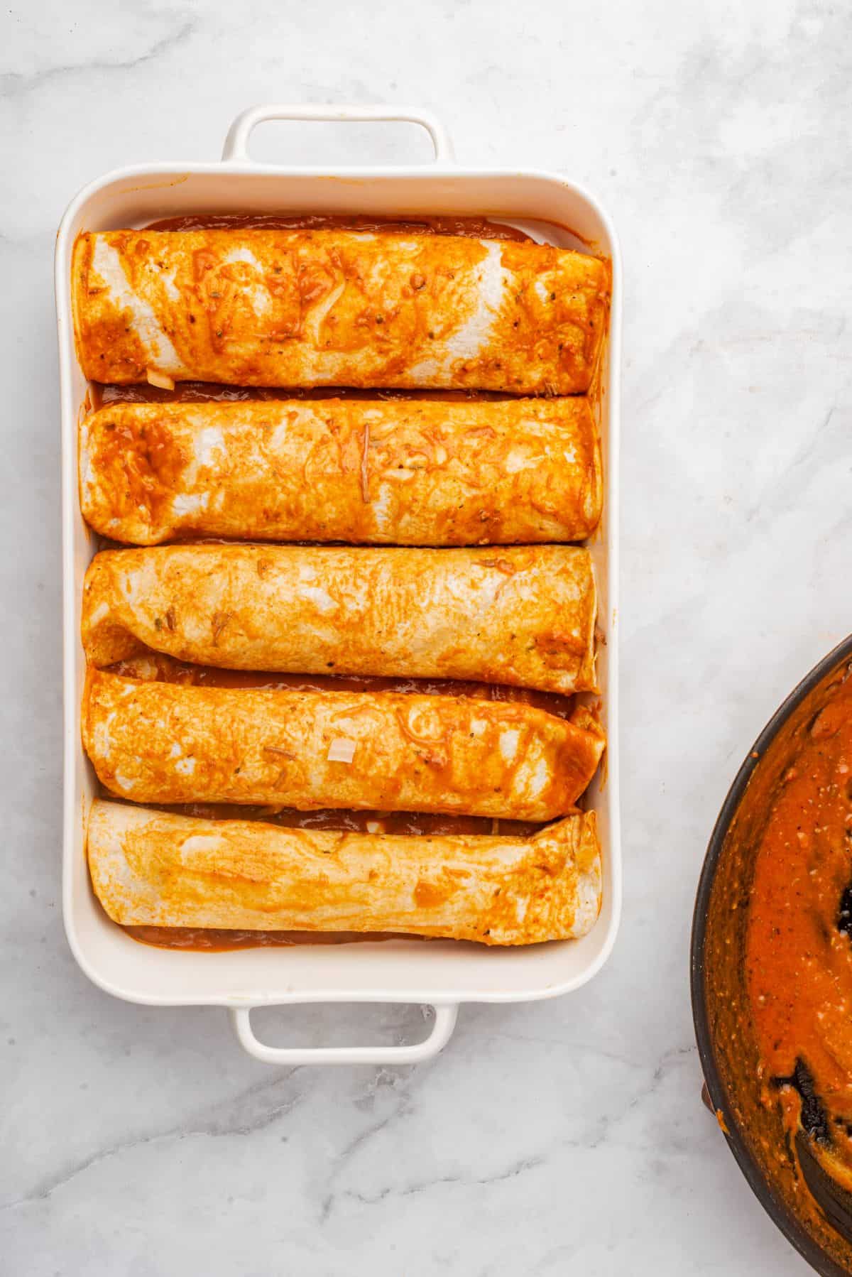 Tortillas coated in red sauce are rolled and placed in a baking dish.