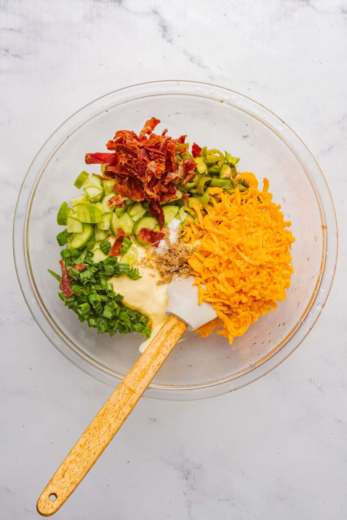 A large mixing bowl filled with mayonnaise, jalapenos, cheddar cheese, bacon, pickles, and scallions with a spatula.