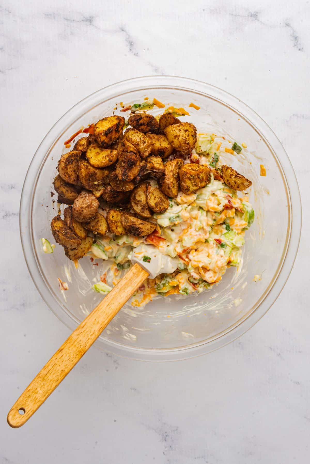 Roasted potatoes in a large mixing bowl with mayonnaise, jalapenos, bacon, cheddar cheese, pickles, and green onions.