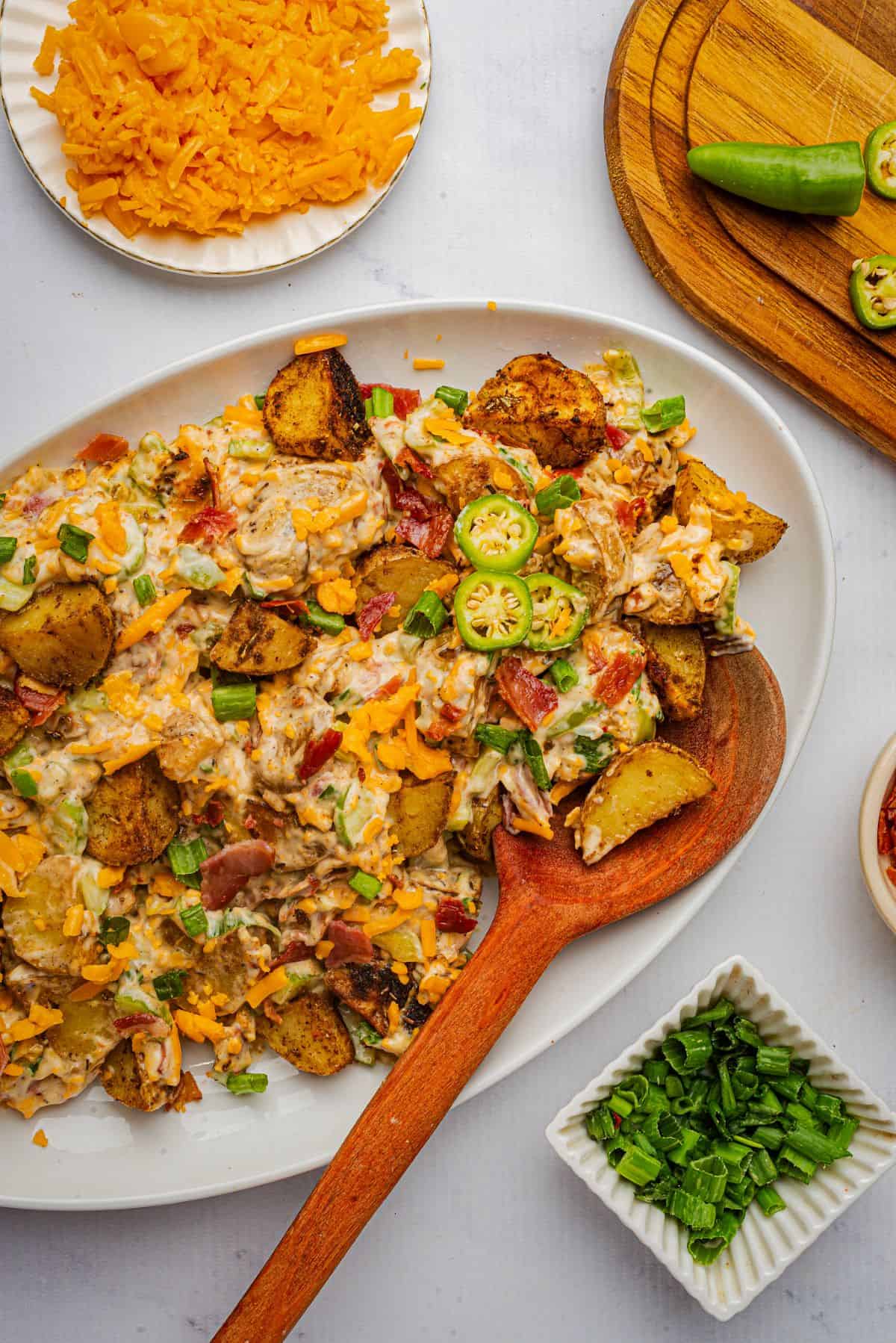 A creamy jalapeno popper potato salad with bacon, cheese, jalapenos, and roasted potatoes on a platter with a wooden spoon.