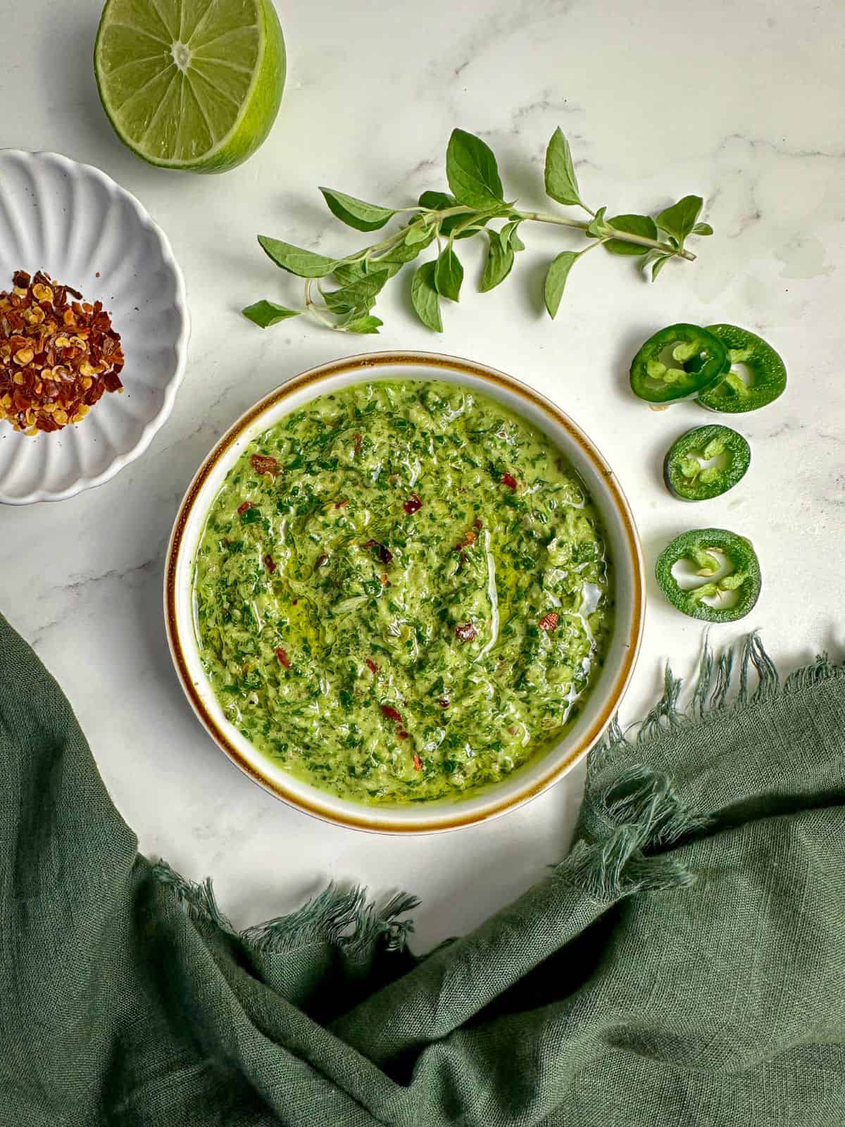 Jalapeno cilantro chimichurri in a bowl surrounded by fresh jalapenos and red pepper flakes.