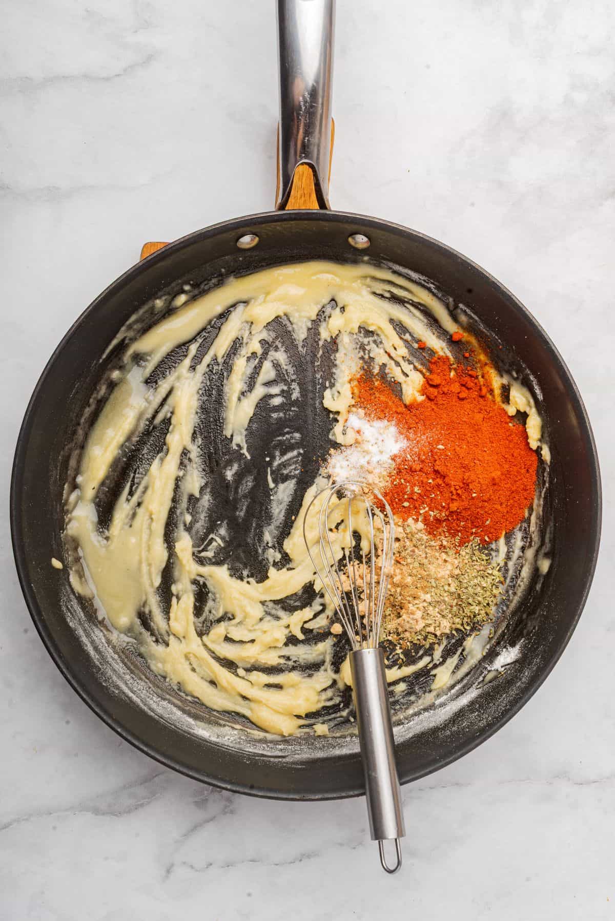 A roux paste has formed in a saucepan and chili powder and other spices have been stirred in with a whisk.