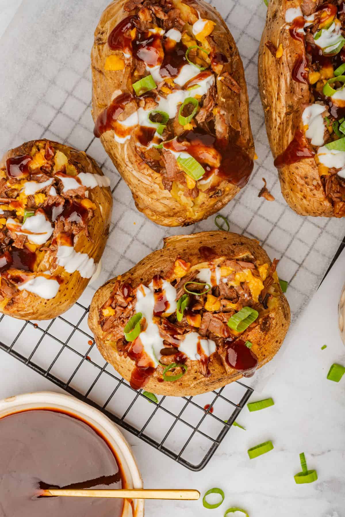 Four loaded brisket baked potatoes on a cooling rack with BBQ sauce, cheese, and sour cream.