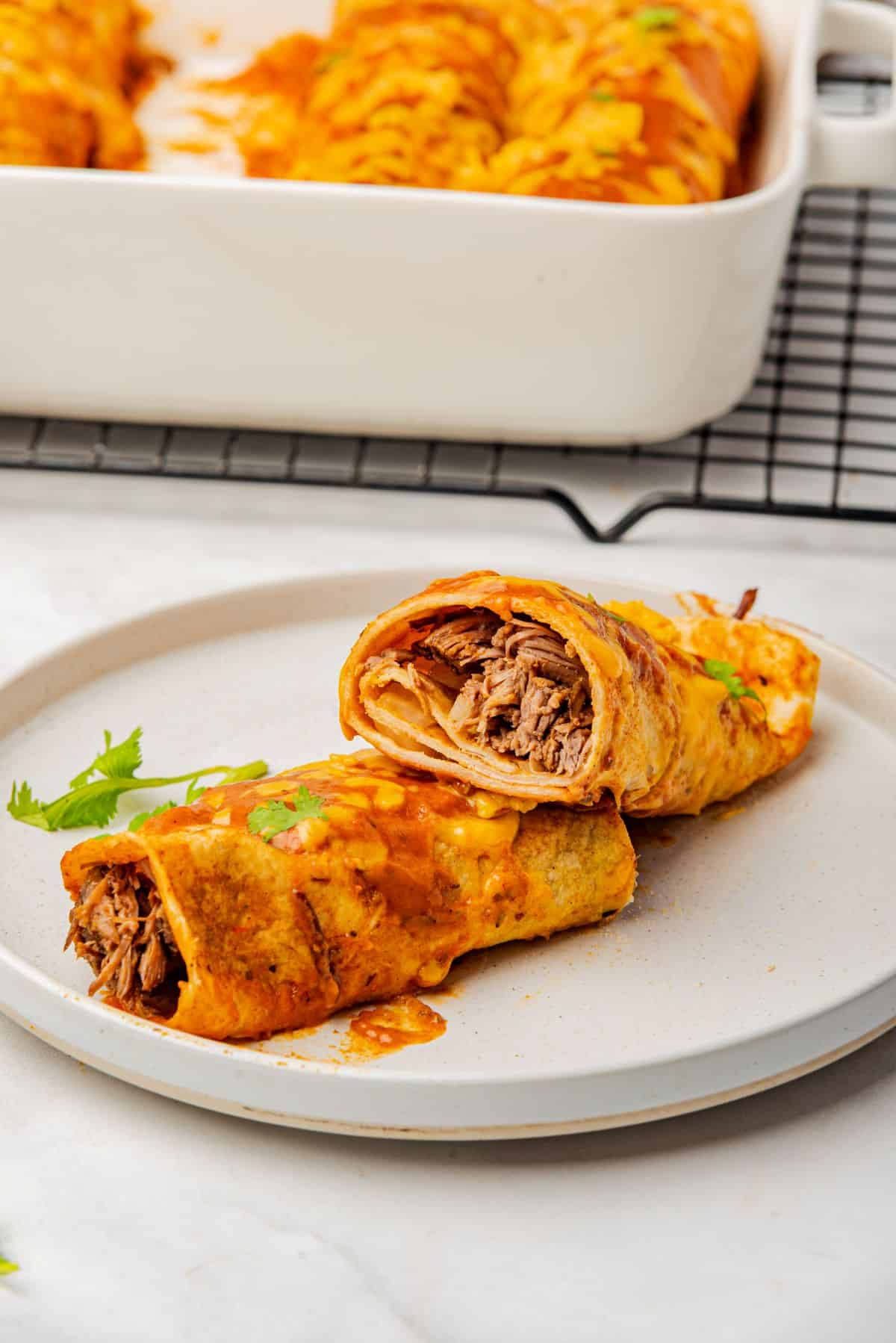 Beef brisket enchiladas with red sauce on a plate cut open to see shredded brisket on this inside of the rolled tortilla.