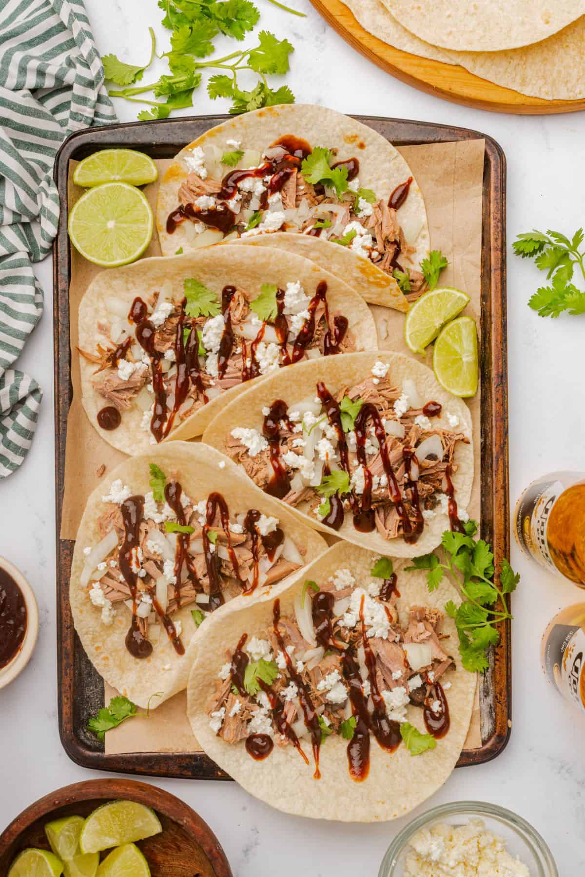 Braised brisket tacos on a sheet pan with toppings of BBQ sauce, onions, cilantro, and cheese.