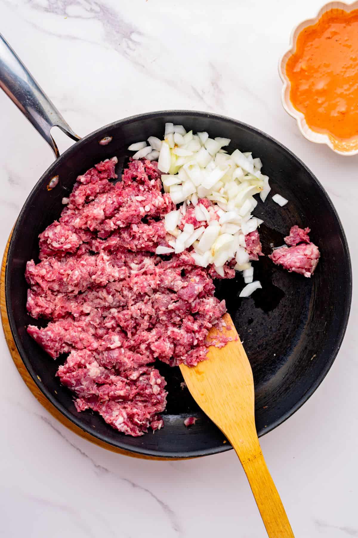 Ground beef and chopped onions in a skillet with a wooden spoon.