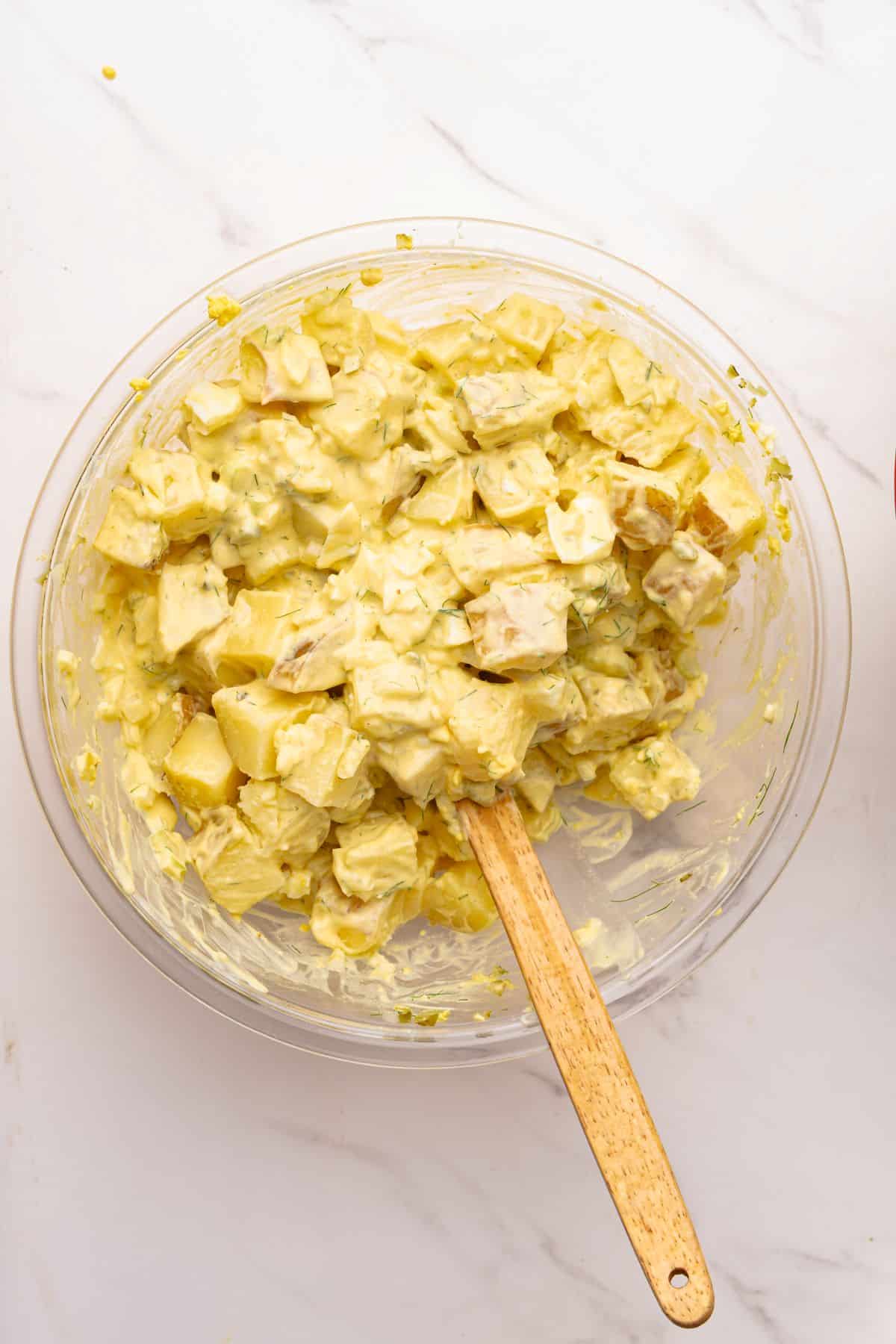 Potato salad with mustard and mayo tossed in a mixing bowl with a wooden spoon.