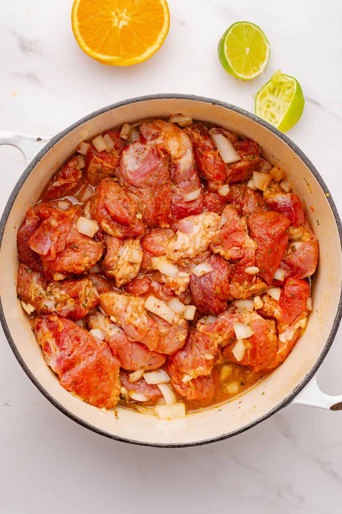 Seasoned pork shoulder chunks in a Dutch oven with chopped onion, minced garlic, orange juice, lime juice, and chicken broth.