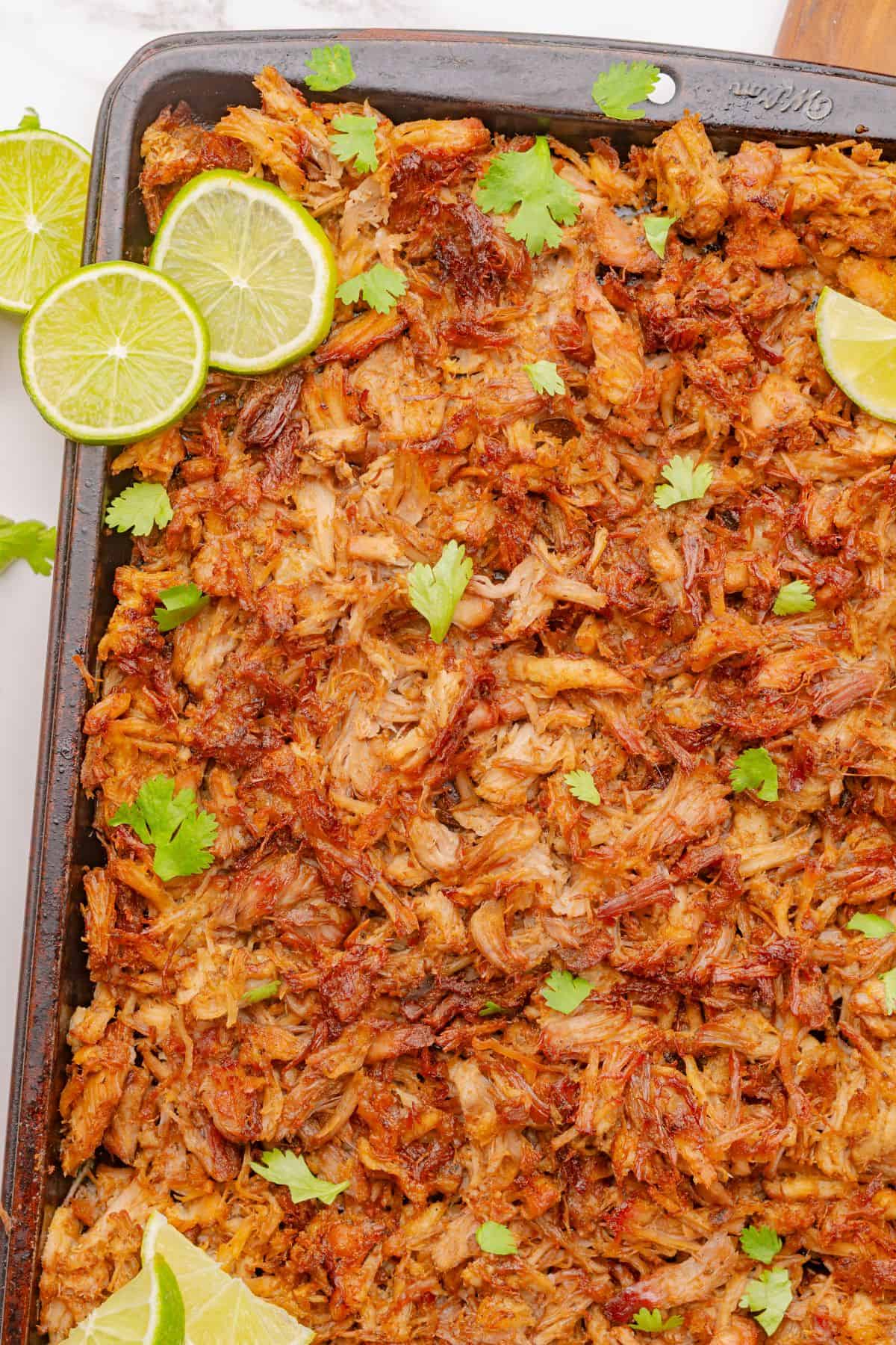 Shredded crispy pork carnitas cooked in a Dutch oven are on a sheet pan with lime slices.