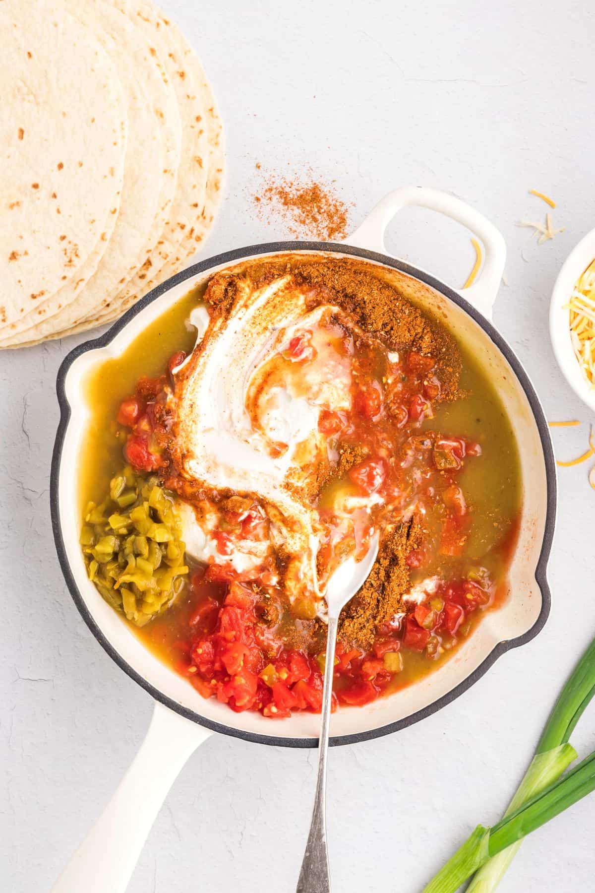 Cream cheese, green enchilada sauce, Rotel, sour cream, green chiles, and chicken stock in a skillet with a spoon.