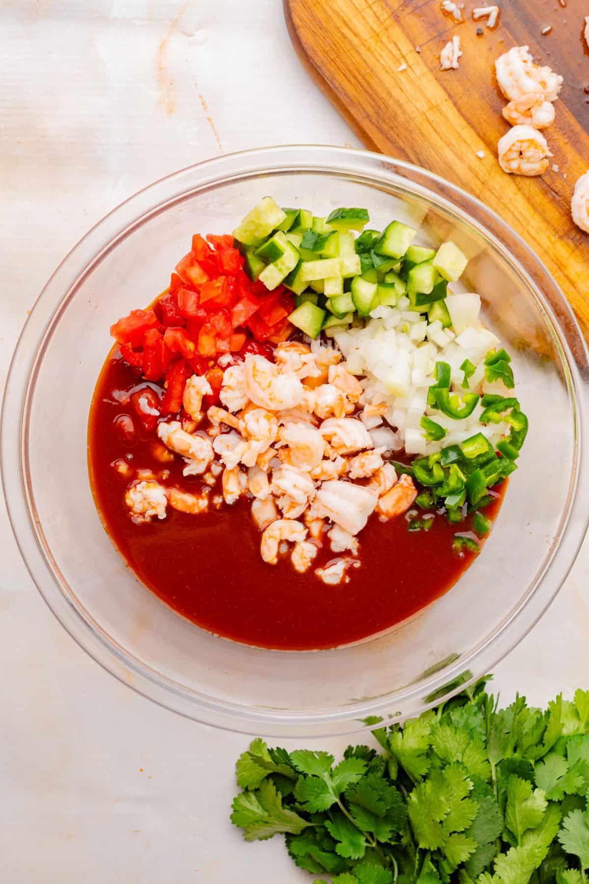 Diced tomatoes, cucumbers, onions, serrano peppers, and shrimp in a mixing bowl with Clamato shrimp cocktail sauce.