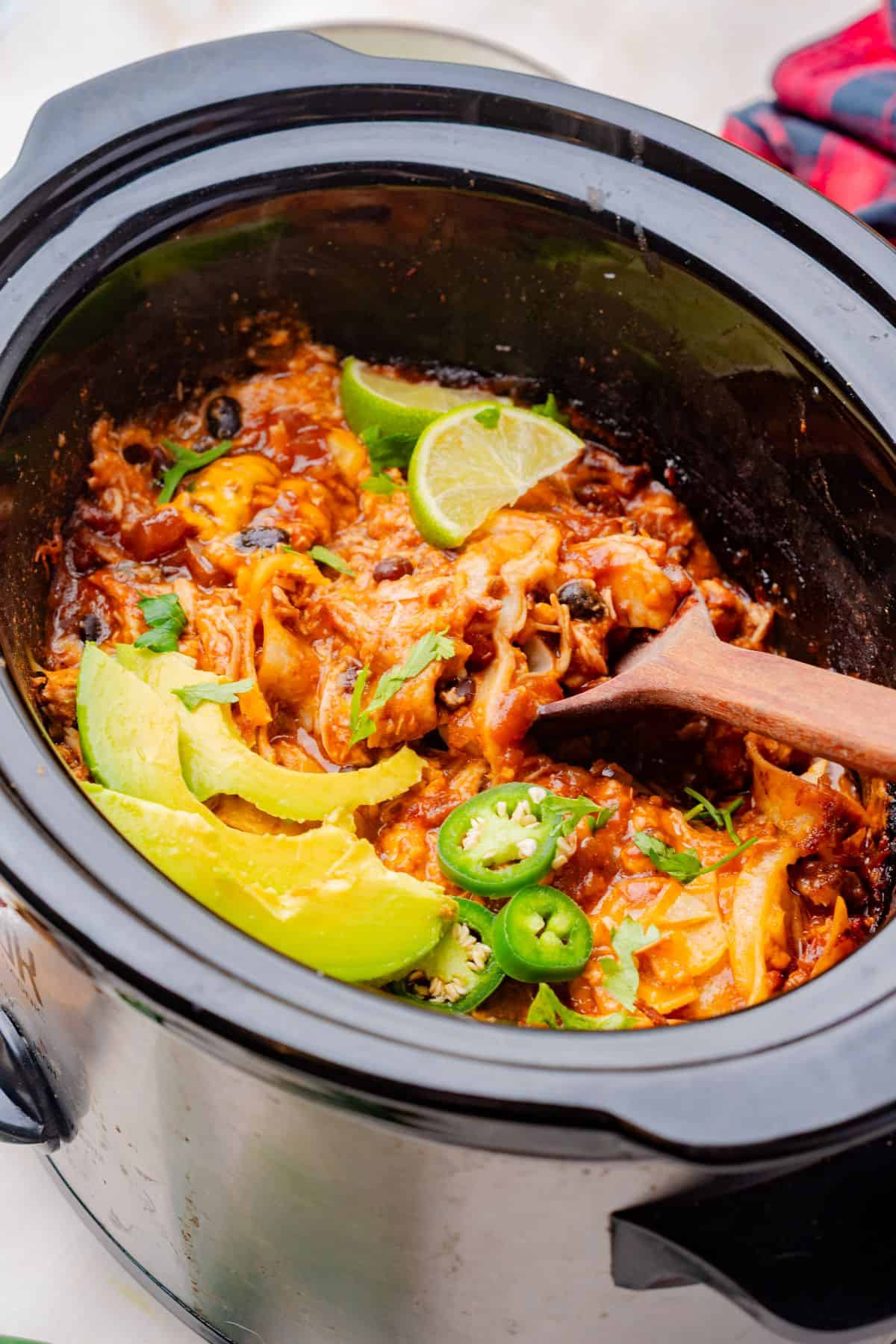 Chicken enchiladas in a crockpot topped with sliced avocado with a wooden spoon.